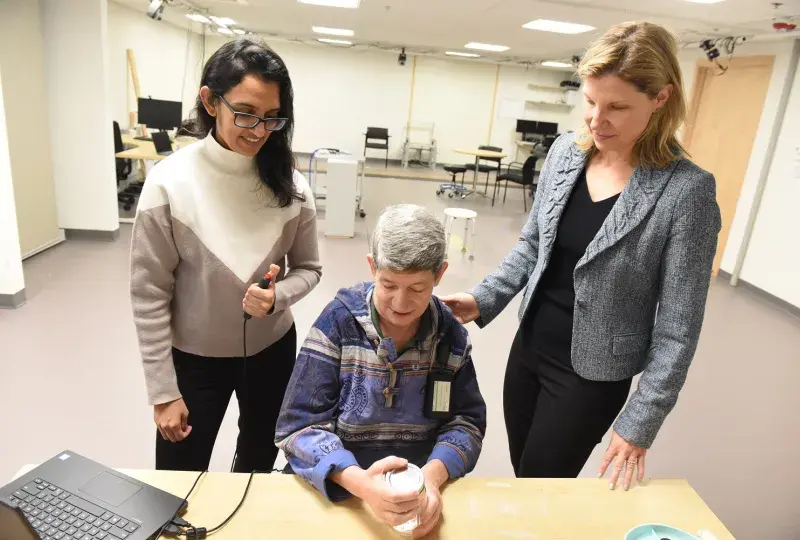two women stand behind a person sitting at a desk one woman has her hand on the shoulder of the woman sitting down