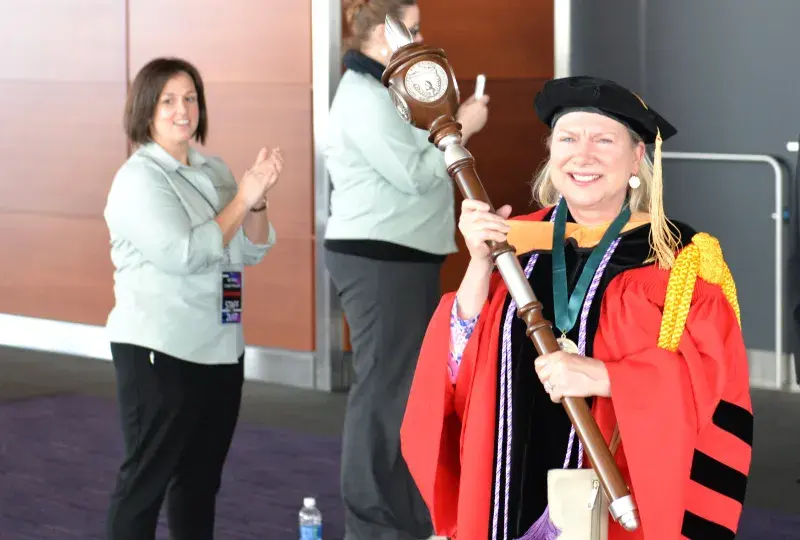 A woman in commencement attire holding a large scepter 