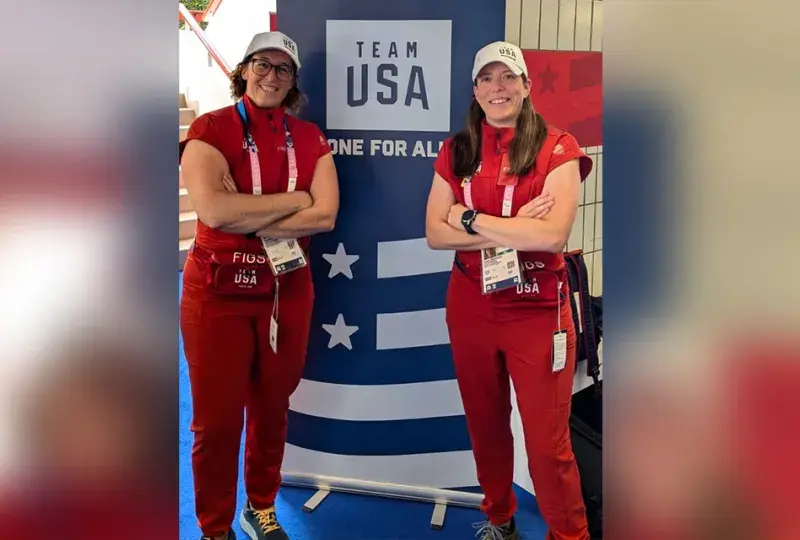 two women in red outfits pose in front of a team USA sign