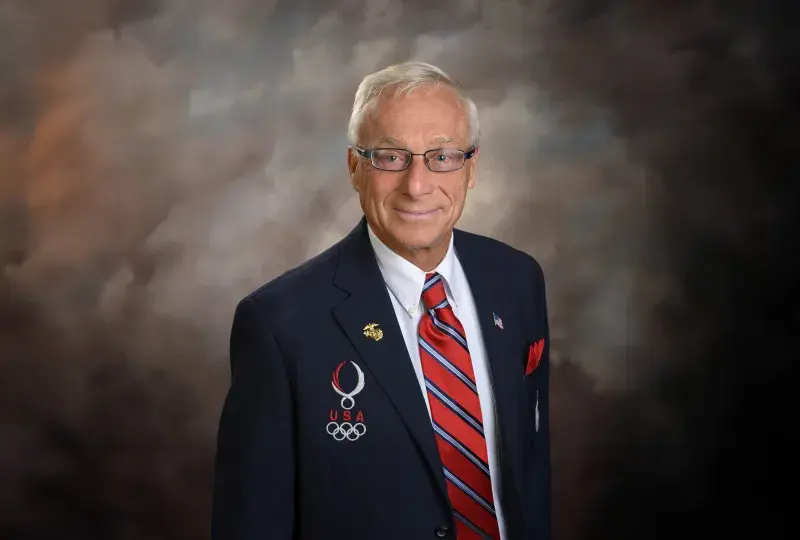 george wears a navy blazer with white Olympic rings on it and a red white and blue striped tie