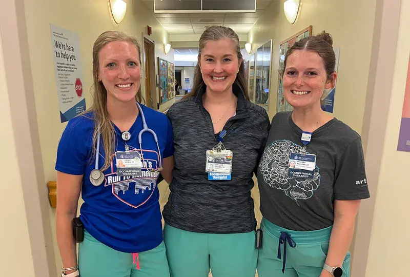 three women in matching scrub bottoms stand in a clinical hallway arm and arm 