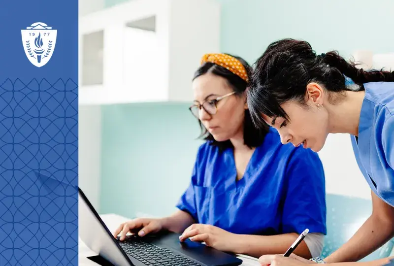 two women in scrubs work at a table with a laptop and pencil