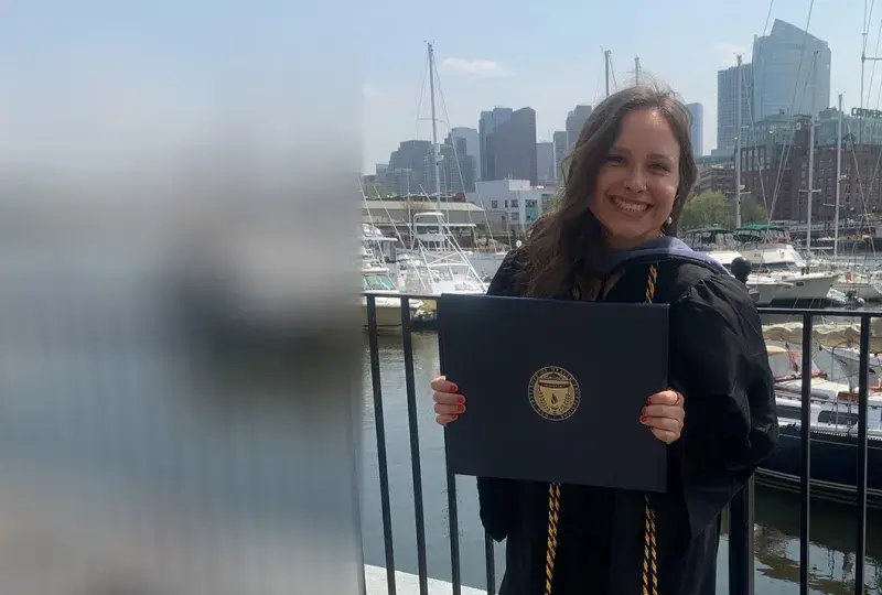 A woman wearing a graduation gown holds a diploma in front of a background of sailboats
