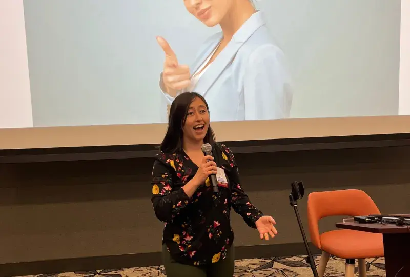 A woman stands in front of a screen holding a microphone