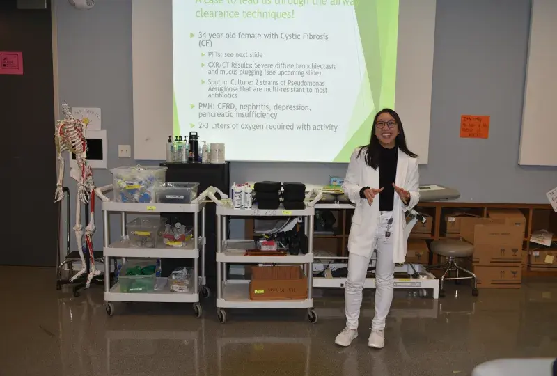 A woman in a white sweater and pants stands in front of a screen in a classroom with medical items behind her