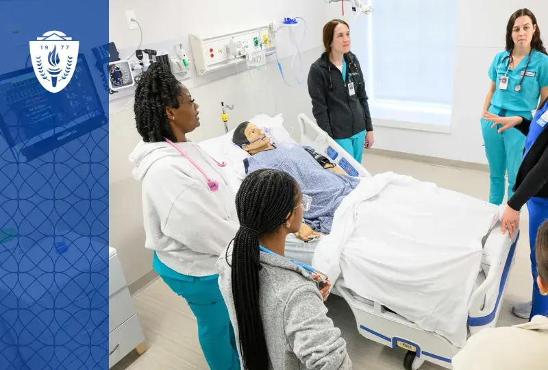 Education in the Health Professions is changing, here we see a faculty member in scrubs surrounded by five students in scrubs around a hospital bed with a manikin