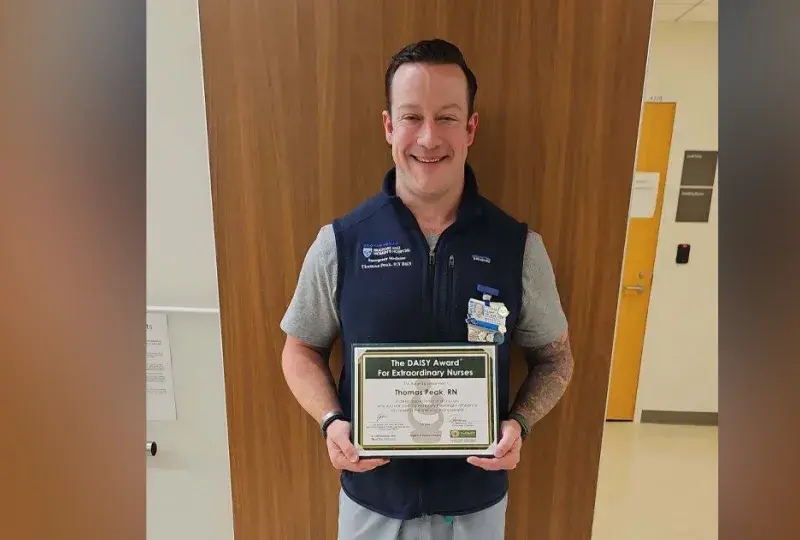 A man stands with his back to a wall holding a certificate for the Daisy Award for Extraordinary Nurses