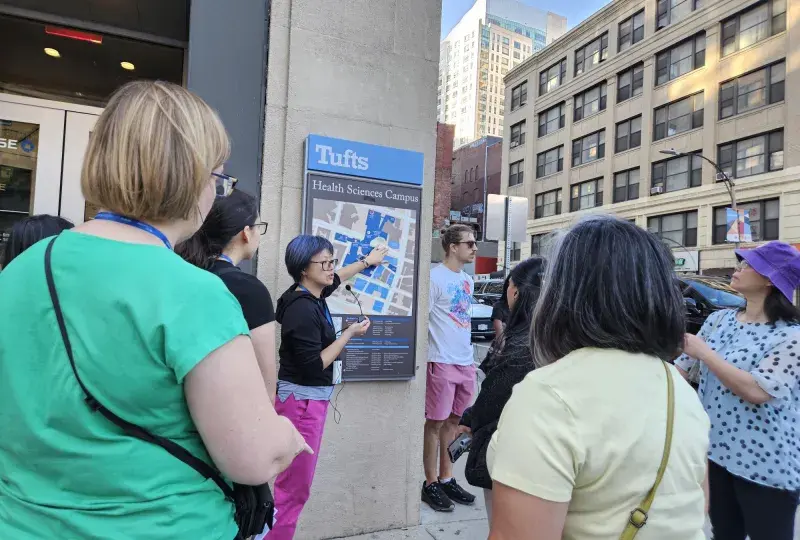 A group of people listen to a woman pointing to a map on a building under a sign that says Tufts