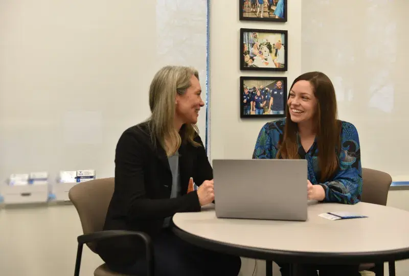 Two women sit at a desk talking with a computer between them