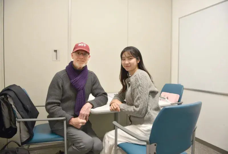 A man in a red baseball cap sits at a table facing the camera while a woman sits next to him facing the camera