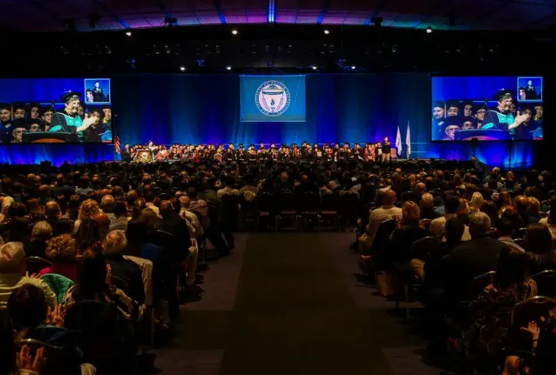 An audience sits in front of a stage while people wearing academic robes sits on the stage
