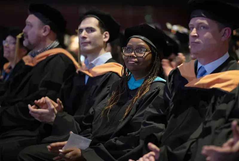 A woman wearing graduation robes sits in a crowd of people 
