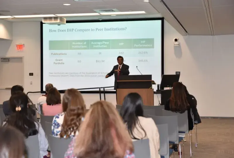 A man stands at a podium with a screen behind him that says "How does IHP Compare to Peer Institutions