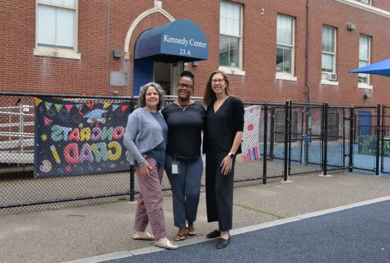 Three women pose outside a building that has a sign that reads Kennedy Center