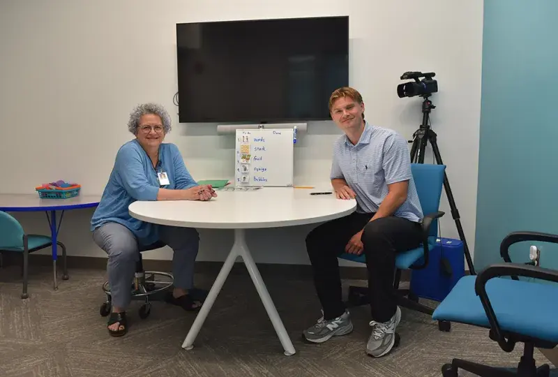 Two people sit at a table with a tv screen behind them 