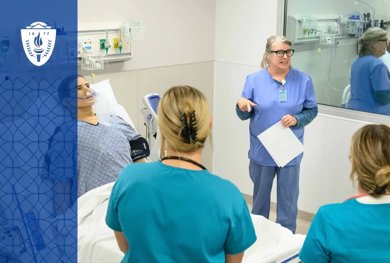 Professor and students wearing scrubs standing around a simulation hospital bed with a mannequin laying in the bed