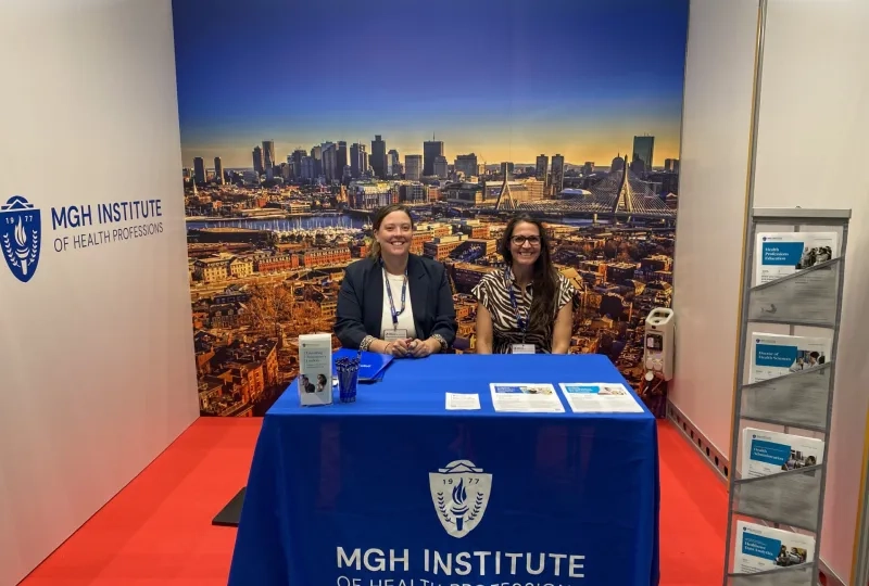 two women sitting at MGH Institute booth with a background of Boston