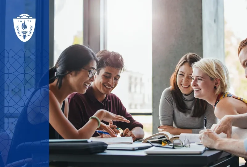 Group of students sitting around desk smiling, reading books, and taking notes.