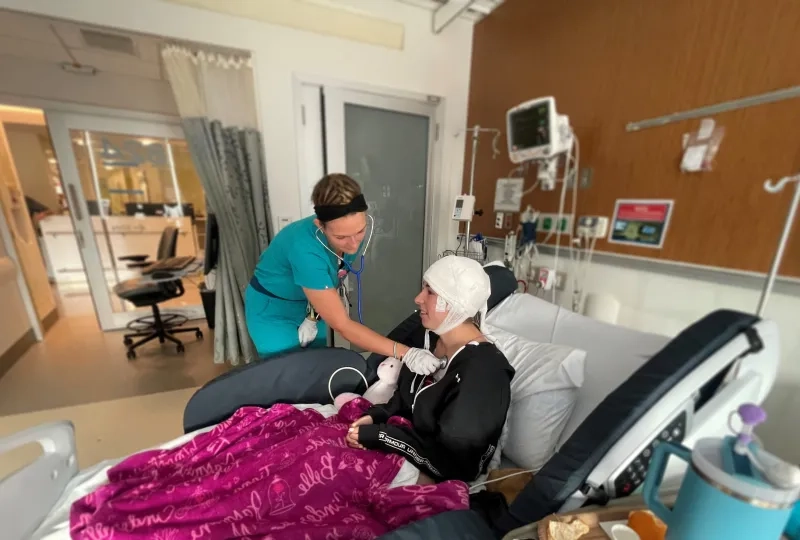 woman in scrubs holds a stethoscope to a woman's chest in a hospital bed