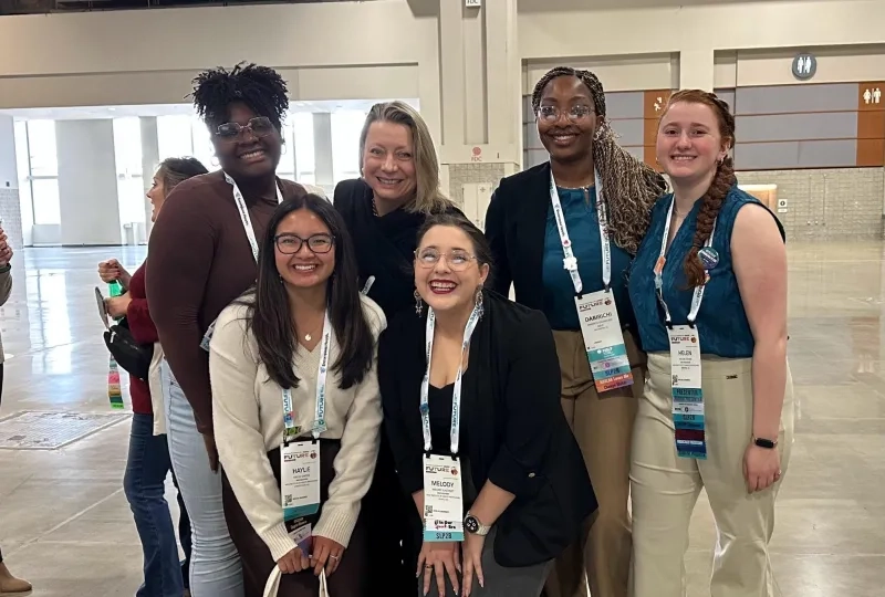 Five women pose in a convention center wearing lanyards