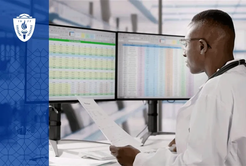 Man wearing lab coat sitting a desk and looking at spreadsheets on two computer monitors