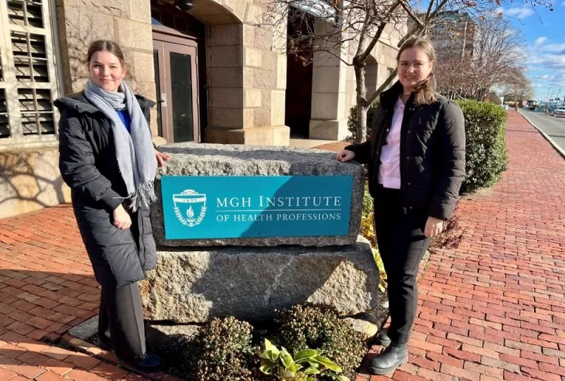 Two women pose on either side of a rock with sign for MGH Institute