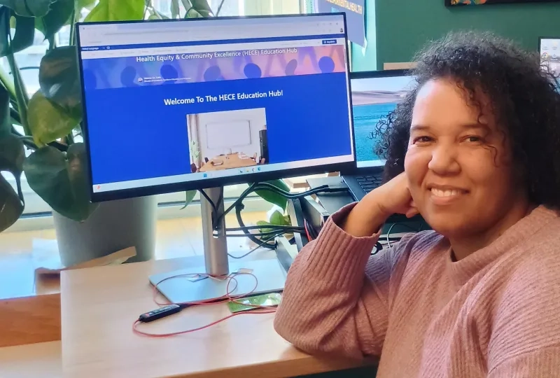 A woman looks at the camera with a computer screen behind her
