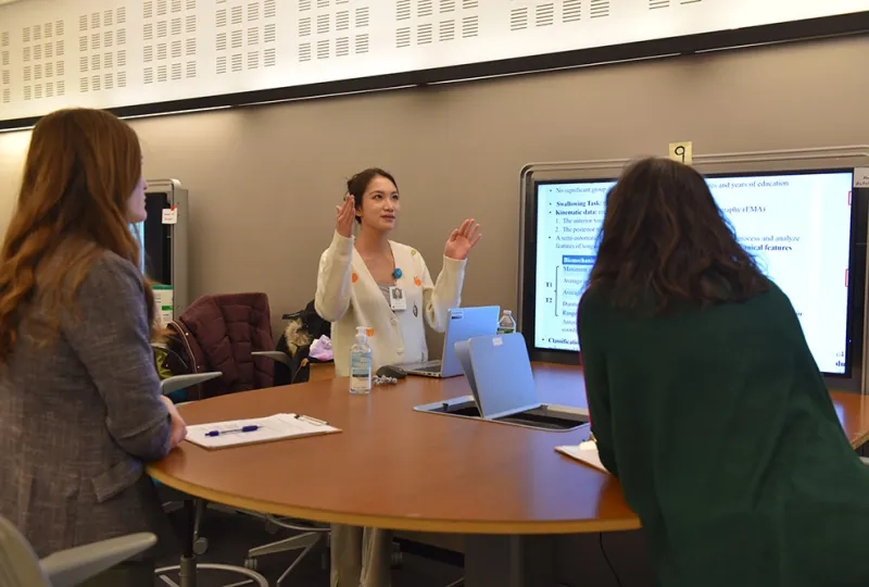 A woman stands in front of screen and talks to two people facing her