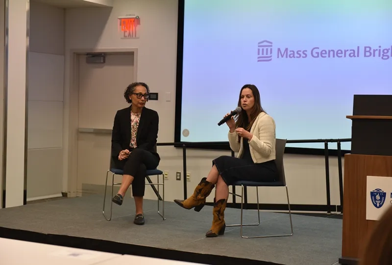 Two women sit on a stage with one speaking into a microphone