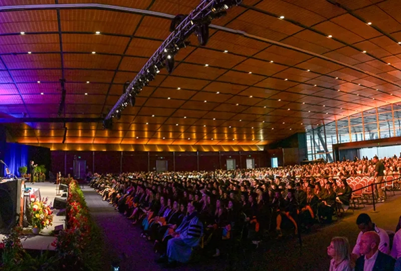 A large room with people dressed for commencement and an audience behind them