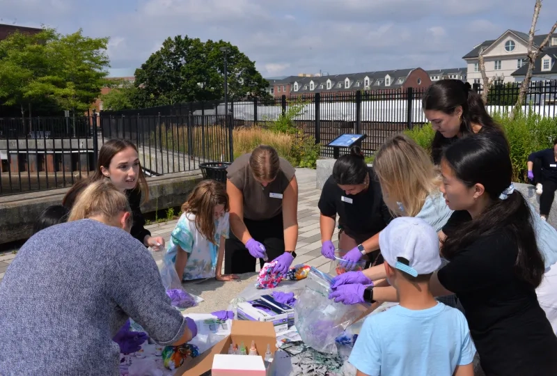 Kids and adult wear purple gloves and stand around a table dying shirts