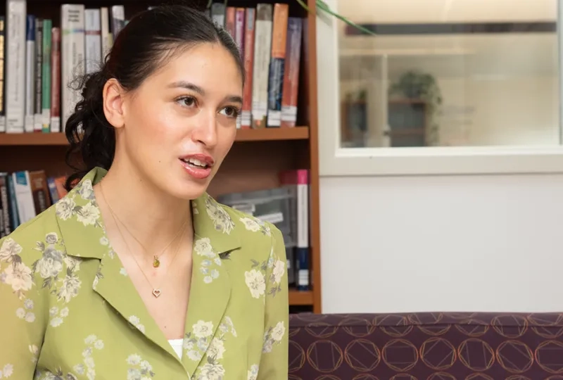 Nicole Vasquez sits on a couch with a bookcase behind her talking to someone off camera