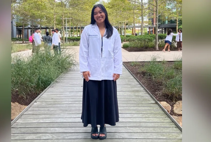 Ashley Ma stands on a walkway with landscaping on the side and trees in the background