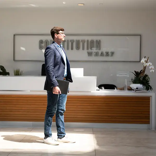 Man holding a laptop at his side stands in front of a desk in a shaft of sunlight