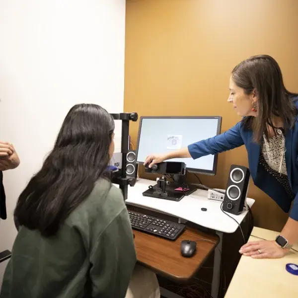 woman rests chin on black device and looks at a screen and camera while a woman points at the screen