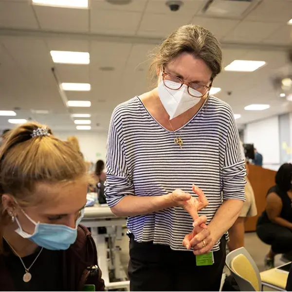 A faculty member wearing a facemask and glasses lists items using her fingers to count and looks over the shoulder of a student wearing a facemask