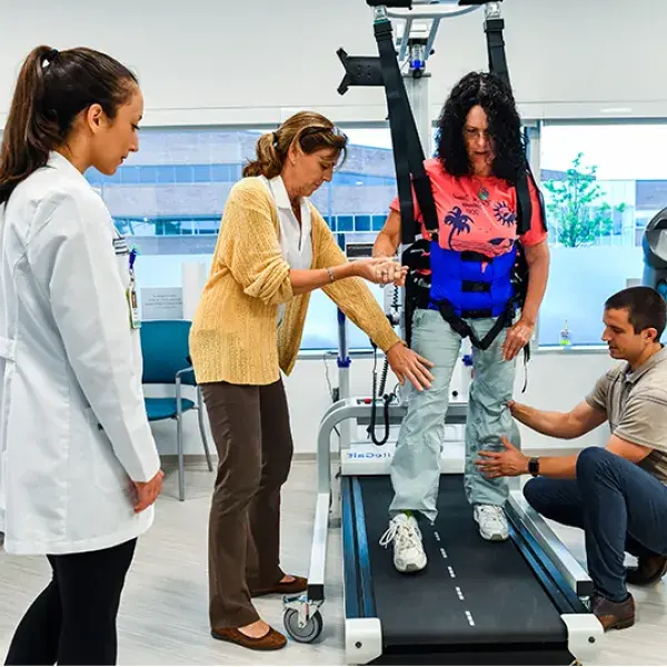 a woman in a harness walks on a treadmill helped by staff members