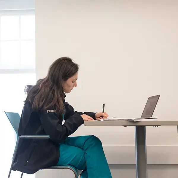 student in scrubs sits in front of a laptop and writes on paper with a pen