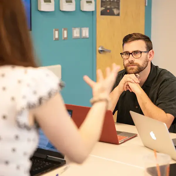 man with glasses rests his chin on his hands attentively as he listens to a teacher