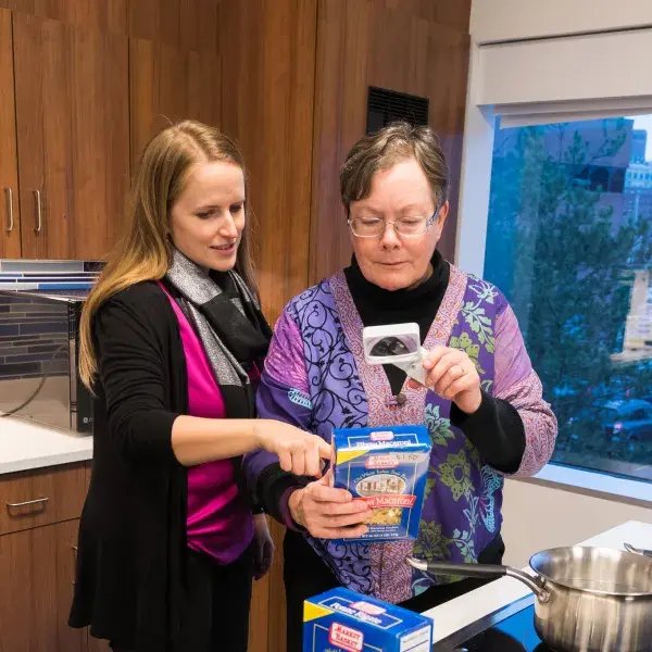 woman points to the back of a pasta box that a second woman holds up to a magnifying glass in front of a stove