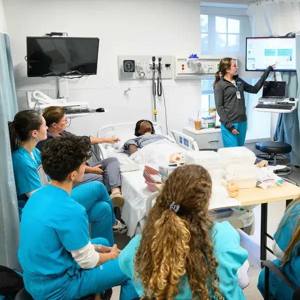 a woman points at a screen in front of other people in scrubs in front of a hospital bed