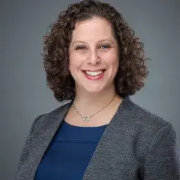 rachel has curly brown hair and wears a grey blazer