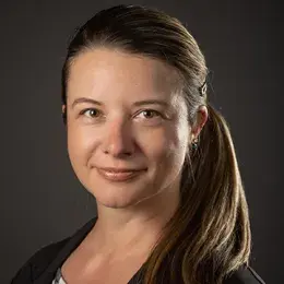 rachel has a long brown ponytail and wears a black jacket over a black and white palmtree print shirt