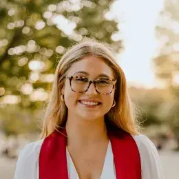 Sydney Zech has blond hair and glasses. She is wearing a white dress with a red graduation ribbon.