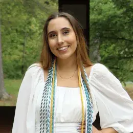 Maegan Mannes is in a white dress with graduation cords and has long brown hair