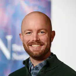 Robert Cavanaugh has a bald head and a brown beard. He is wearing a black sweater over a blue checkered dress shirt.