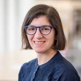 Emily Rosen has short, straight, brown hair, is wearing blue glasses, and is wearing a dark blue blouse.