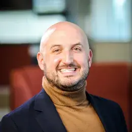 Greg Landry is bald and is wearing a brown turtleneck and navy blazer