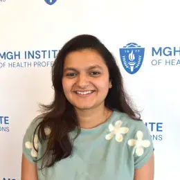 Woman with long black hair wearing a light blue shirt with a flower design on it standing in front of a background featuring the MGHIHP logo