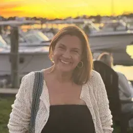 Woman wearing a black shirt and white sweater smiling and looking at camera. There is a sunset and boats on the water in the background.
